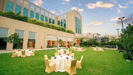 An elegant outdoor event space with round tables set up on a lush green lawn, adjacent to the hotel - Grand Madurai by GRT Hotels