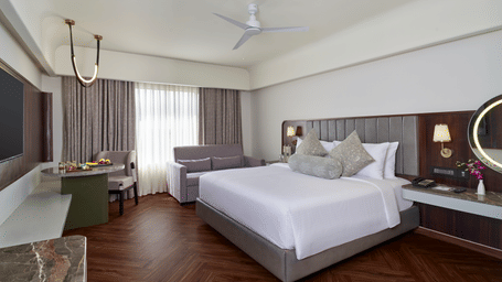 The Elite Room at Swosti Premium, Bhubaneswar, featuring a double bed, bedspreads, pillows,  cushions, a table, large windows covered in curtains, a mirror, a television,  wall mounted lamps and a fan. 