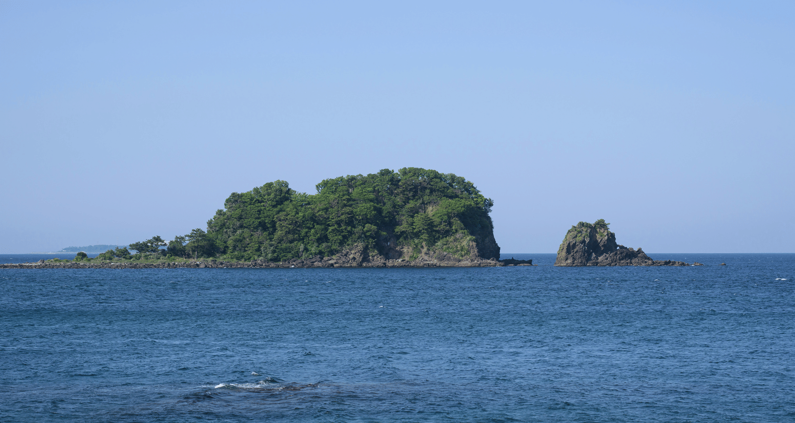 A small, rocky, green island in the middle of a blue ocean under a clear, bright sky.