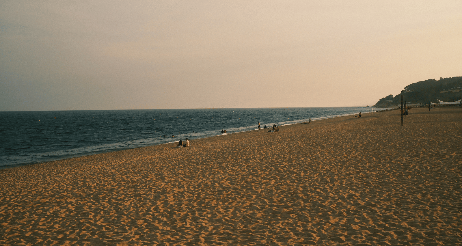 A wide, sandy beach extends under a hazy, pale sky with a few distant figures near the water's edge.