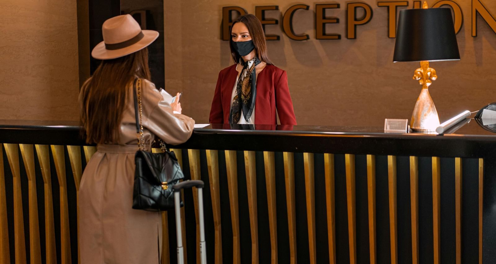 A woman wearing a hat and a light coat at a hotel reception desk, where a receptionist wearing a face mask stands ready to assist her.