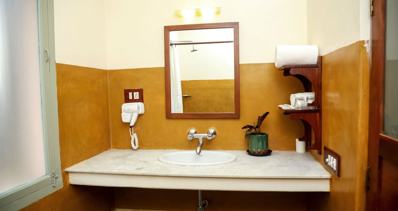 A bathroom with a sink, toiletries, and a mirror on the wall, A sitting area, a cupboard, and a tall lamp | Mango Hill La Serene, Pondicherry