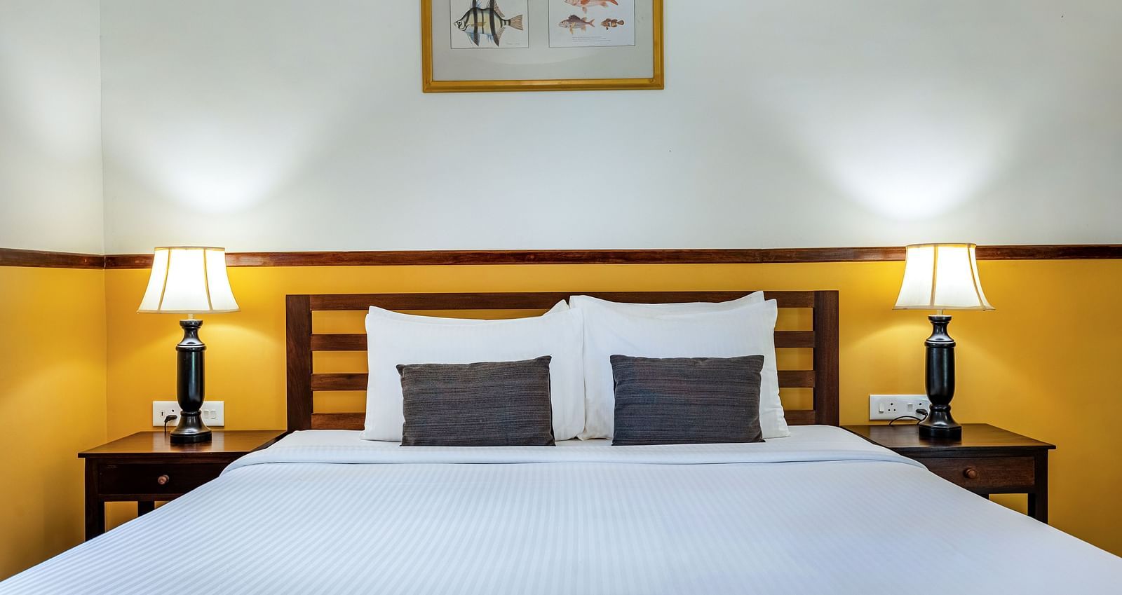 A neatly made double bed with pillows, flanked by matching lamps on nightstands, beneath a framed picture in a room near Pondicherry | Mango Hill Shallow Beach Resort ECR