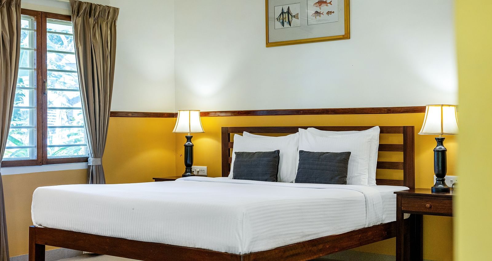 Bedroom featuring a wooden double bed with white linens, flanked by nightstands and lamps, with a framed picture on the wall | Mango Hill Shallow Beach Resort ECR