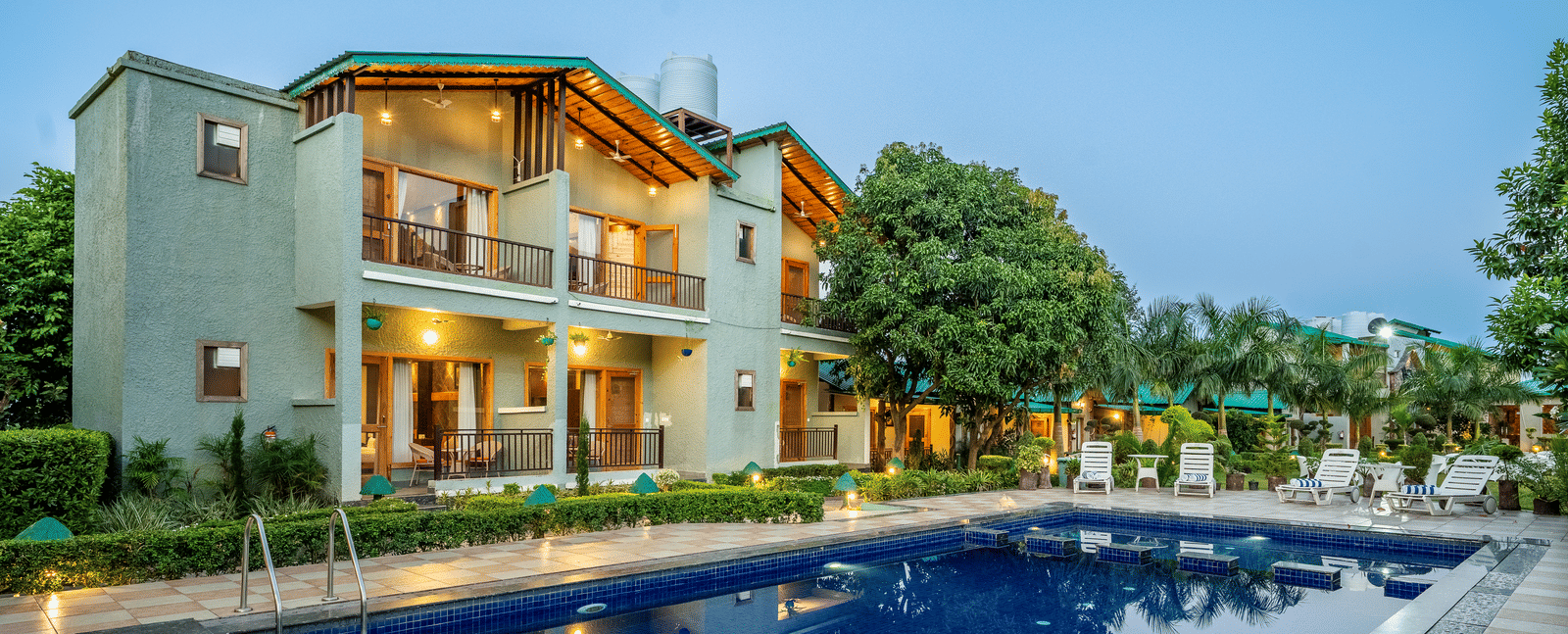 Outdoor swimming pool with tiled deck, lounge chairs, and a two-storey building with balconies and lights turned on, surrounded by trees at Corbett Nirvana Resort.