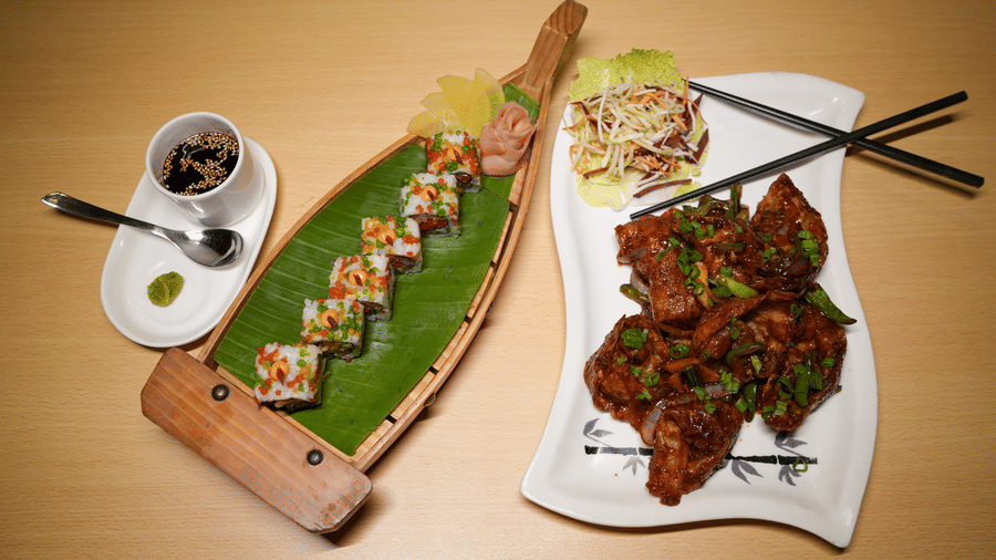An overhead view of an Asian food platter at Park Plaza Ludhiana featuring stir fried dishes sushi on a banana leaf wooden board chopsticks and a dipping sauce bowl.
