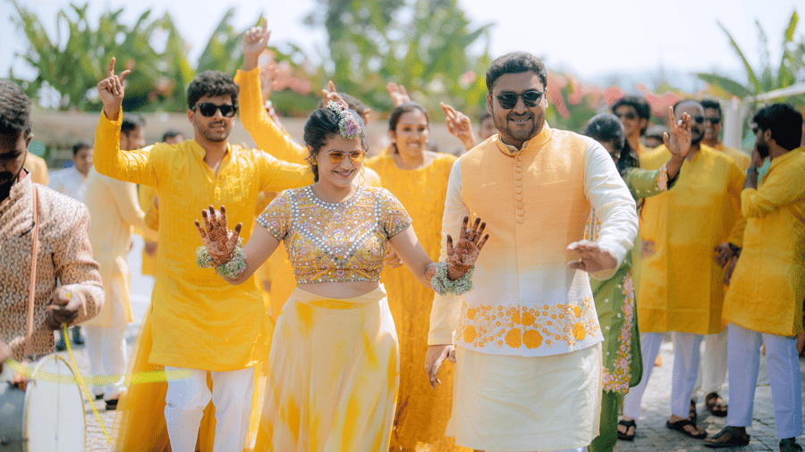 A couple with their friends and family dancing in the background with yellow colour as the dress code at one of the party halls in Coimbatore inside Darza Resorts with clear blue sky in the background.