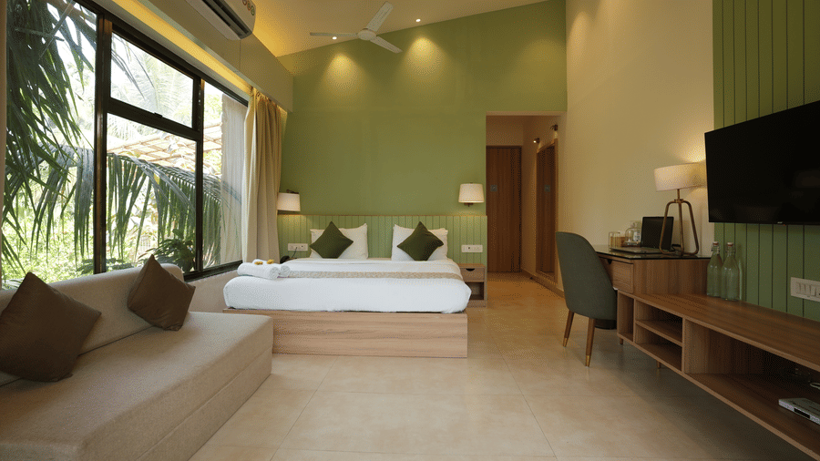 A bedroom at Oceanature Resort, Alibaug with a king size bed against a green wall, a sofa, a long desk, work desk, chair, a wall-mounted television, and large windows.