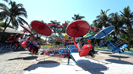 Riders on the Sky Glider in red and blue seats hanging from arms topped with disc-shaped canopies, against palm trees and blue sky at MGM Dizzee World, Chennai.
