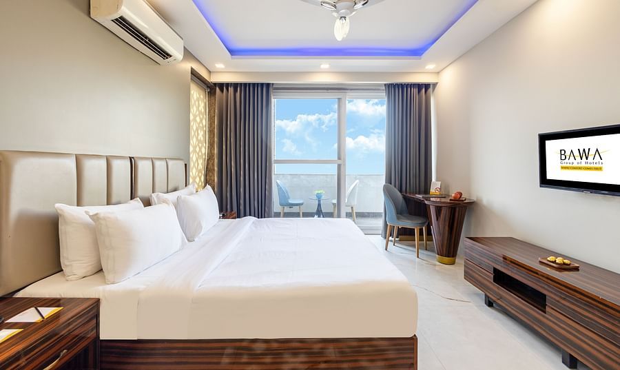 Side view of a king size bed in a room with elegant decor and a balcony view at Hotel Bawa, Gurgaon.