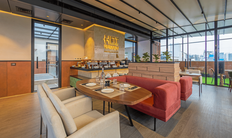 Indoor dining area at Koi Garden restaurant within Hotel Bawa Gurgaon. Tables and chairs are arranged across the room with a buffet counter in the background. Glass walls line the side of the space.