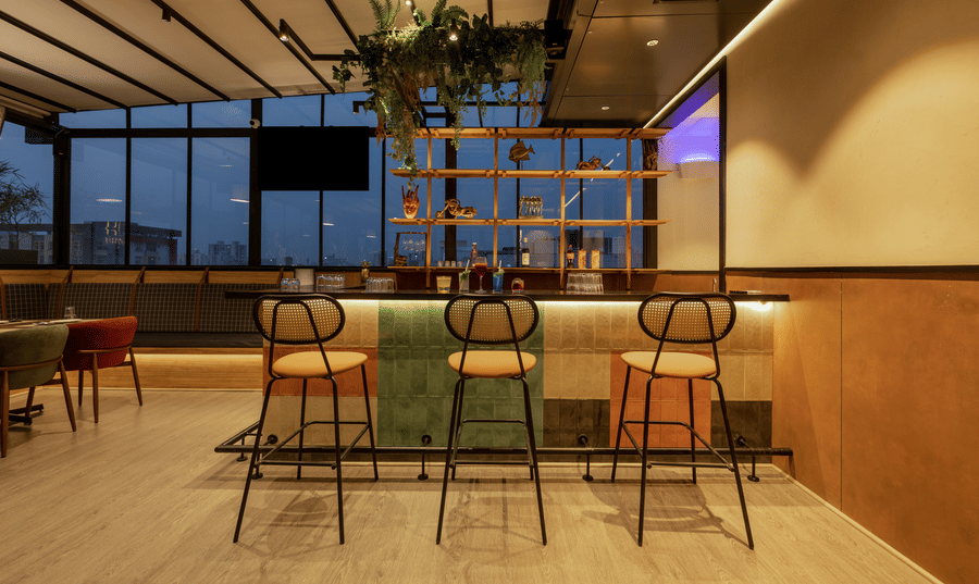 Bar area inside Koi Garden restaurant. Stools are placed along the front of the counter. Shelves with bottles stand beneath the ceiling structure and a hanging plant.