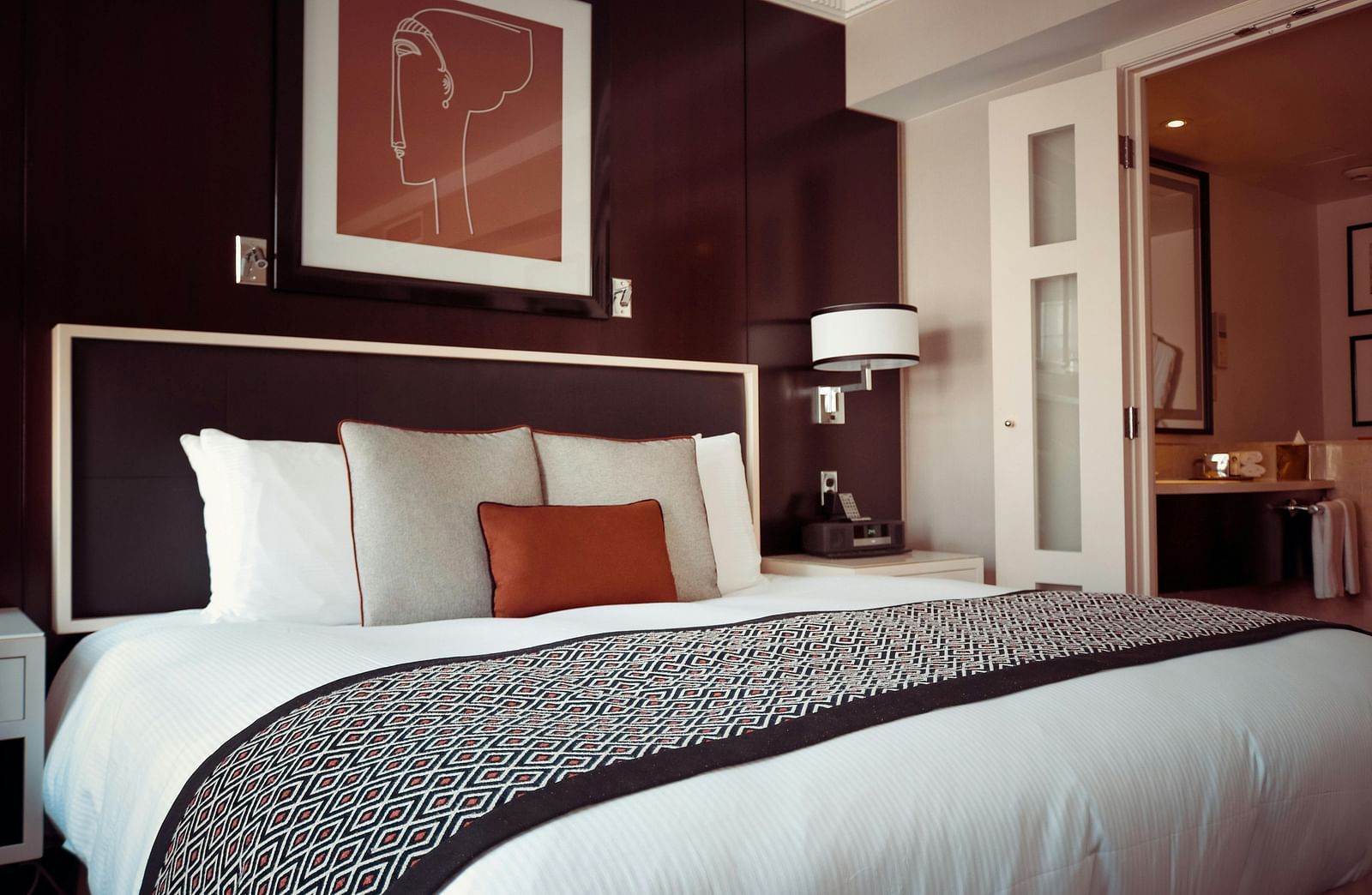 A hotel room displays a dark headboard, white linens, and a pop of colour with an orange pillow.