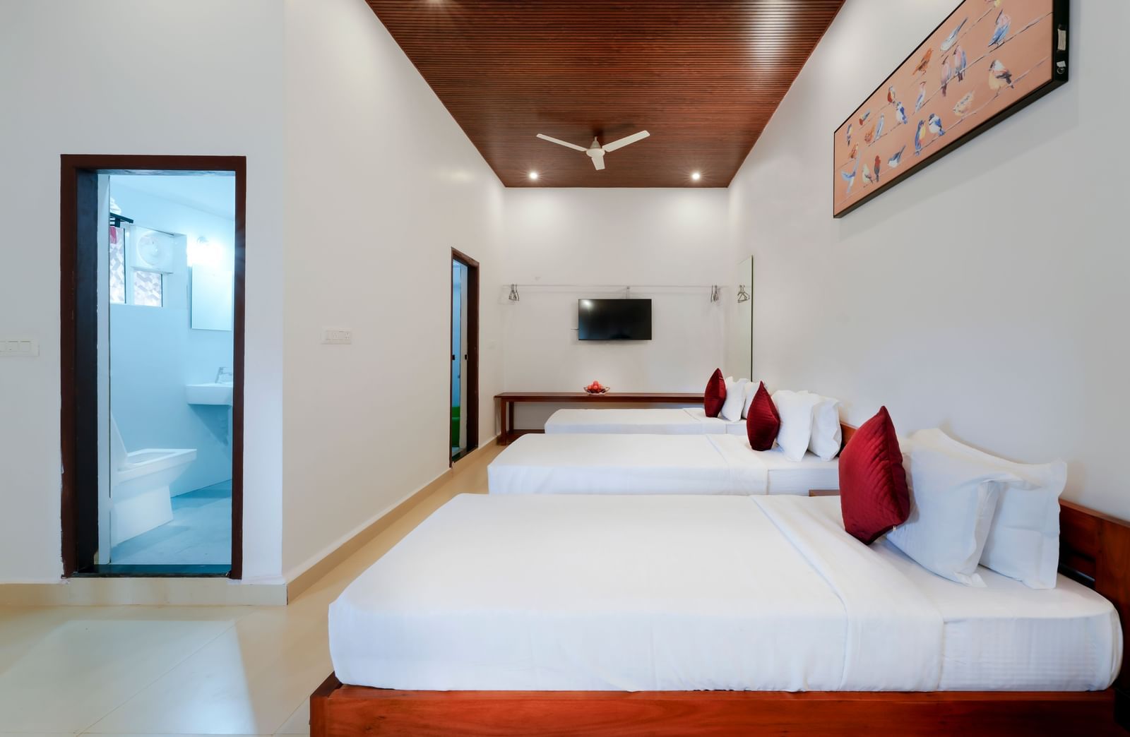 A wide-angle view of the hall of residences at Bhuthanakadu Retreat, featuring four single beds, each with a red pillow, and a bright, minimalist interior with a high wooden ceiling.