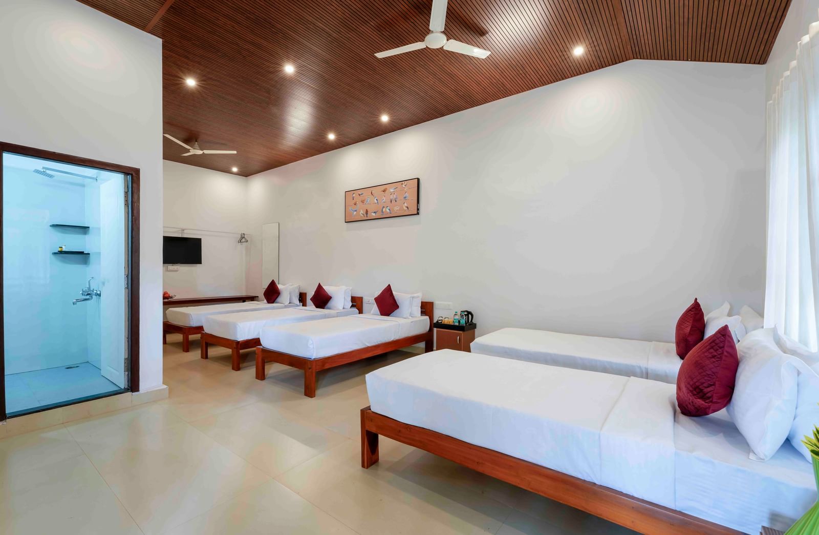 A spacious hall of residences at Bhuthanakadu Retreat, showcasing four single beds with white bedding and red pillows, a modern bathroom, and a warm wooden-paneled ceiling.