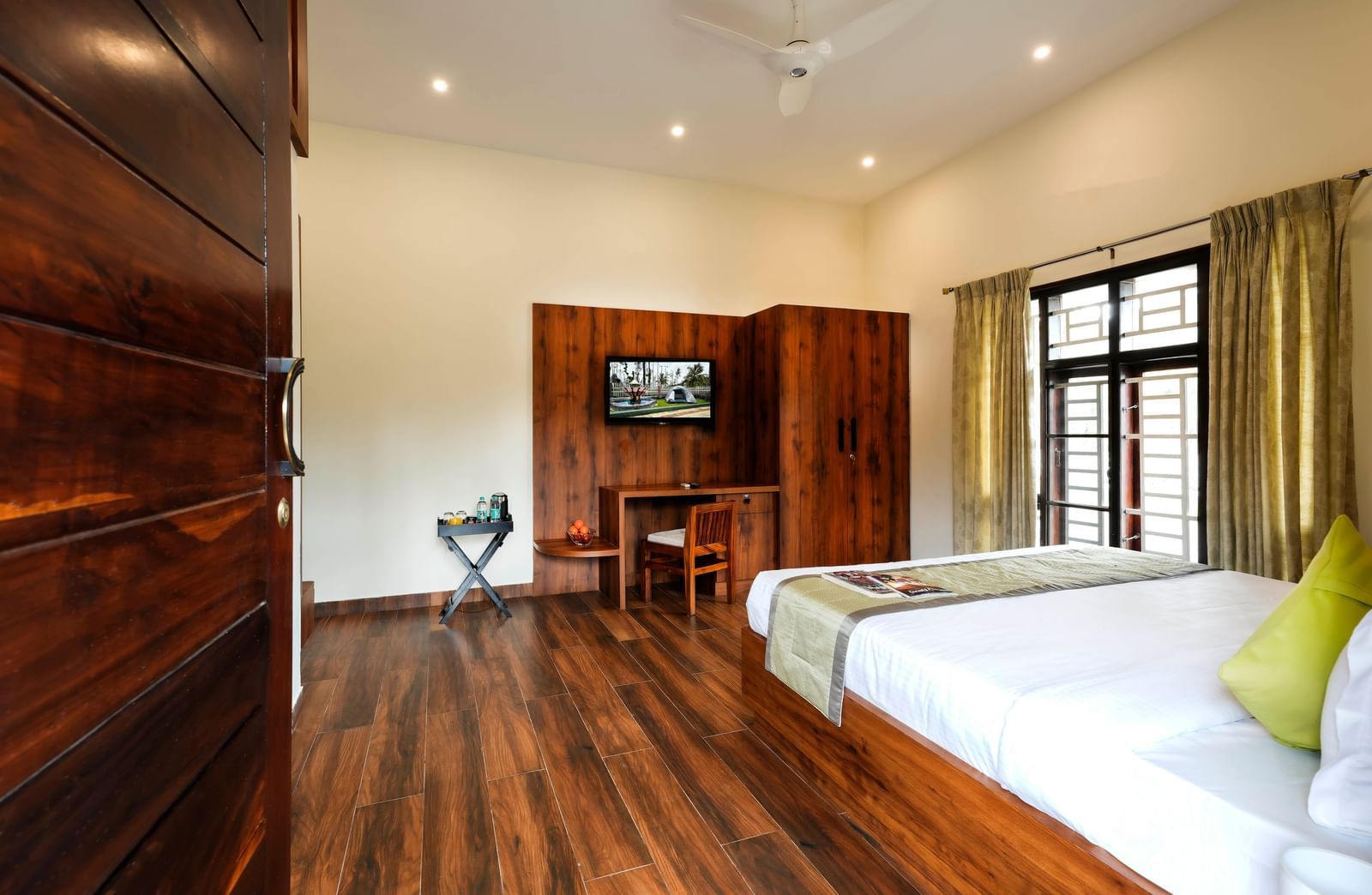 A wide-angle view of the spacious premium room at Bhuthanakadu Retreat, showcasing a large bed, a single bed, a TV on the wall, and rich wooden flooring.