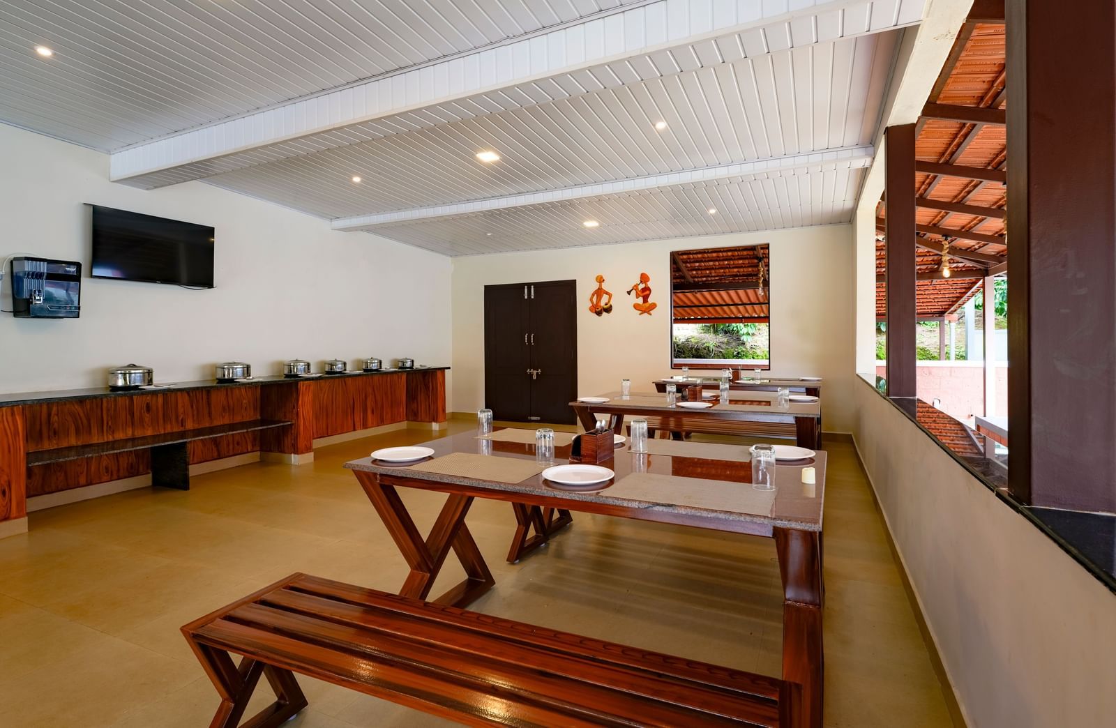 A daytime interior view of the Mane Cafe at Bhuthanakadu Retreat, highlighting a buffet area with covered dishes, a TV on the wall, and long wooden dining tables with benches.