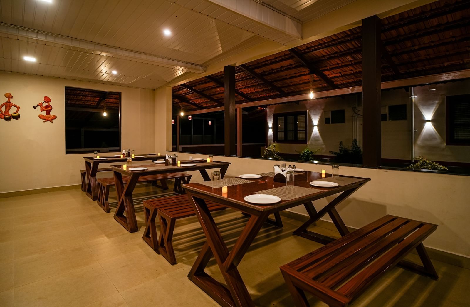 An evening shot of the Mane Cafe at Bhuthanakadu Retreat, showing a well-lit, open-air dining space with rustic wooden tables and bench seating.