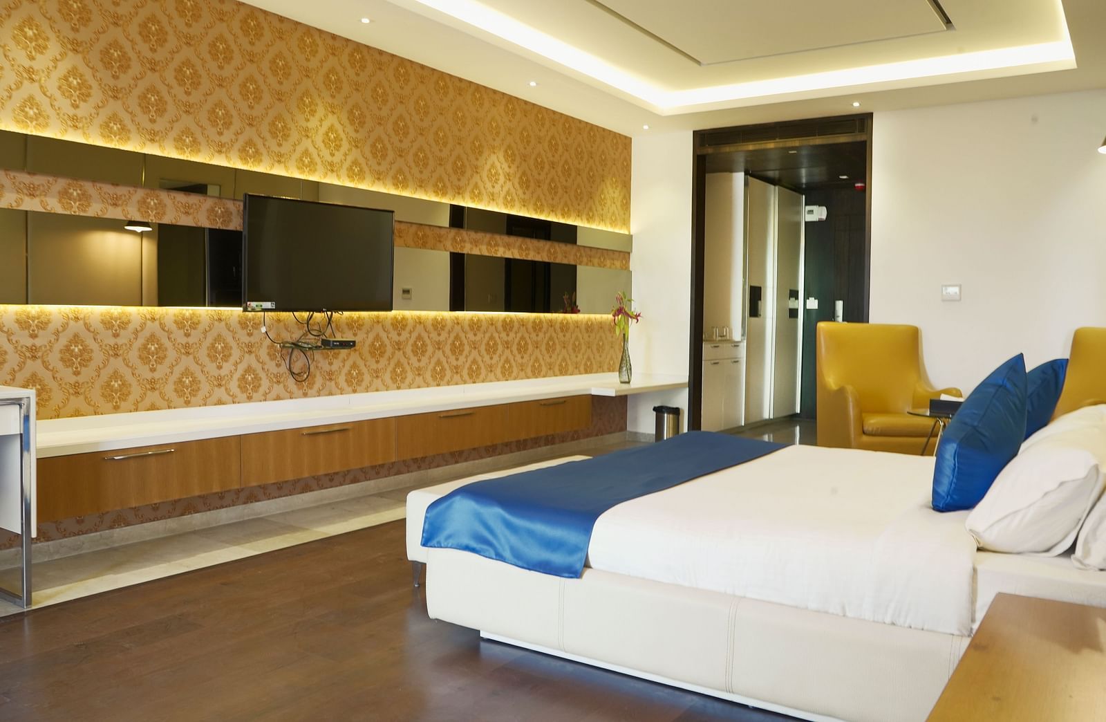 A well-appointed hotel room at BNGV Grandeur Hotel and Banquets, featuring a bed with blue and white linens, a patterned wall, and a dresser with a mirror.