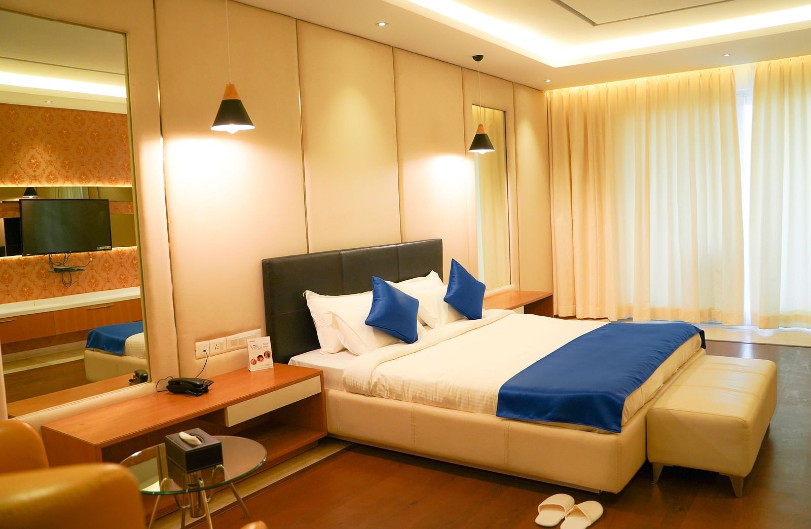 A warm-toned hotel room at BNGV Grandeur Hotel and Banquets, with a bed featuring blue and white linens, and soft lighting creating a cosy ambience.