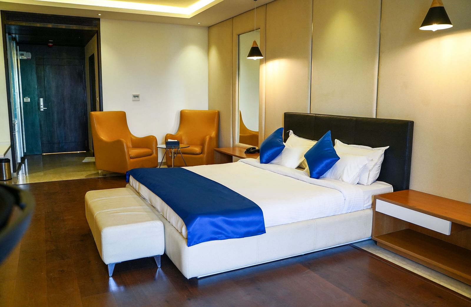 A stylish hotel room at BNGV Grandeur Hotel and Banquets, equipped with a bed featuring a dark headboard and blue accents, complemented by two vibrant orange armchairs.