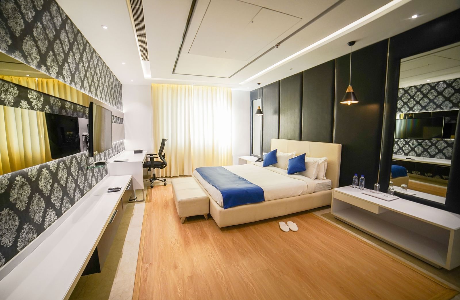 A modern and elegantly designed hotel room at BNGV Grandeur Hotel and Banquets, featuring a comfortable bed with a blue runner, a unique patterned accent wall, and contemporary furnishings.