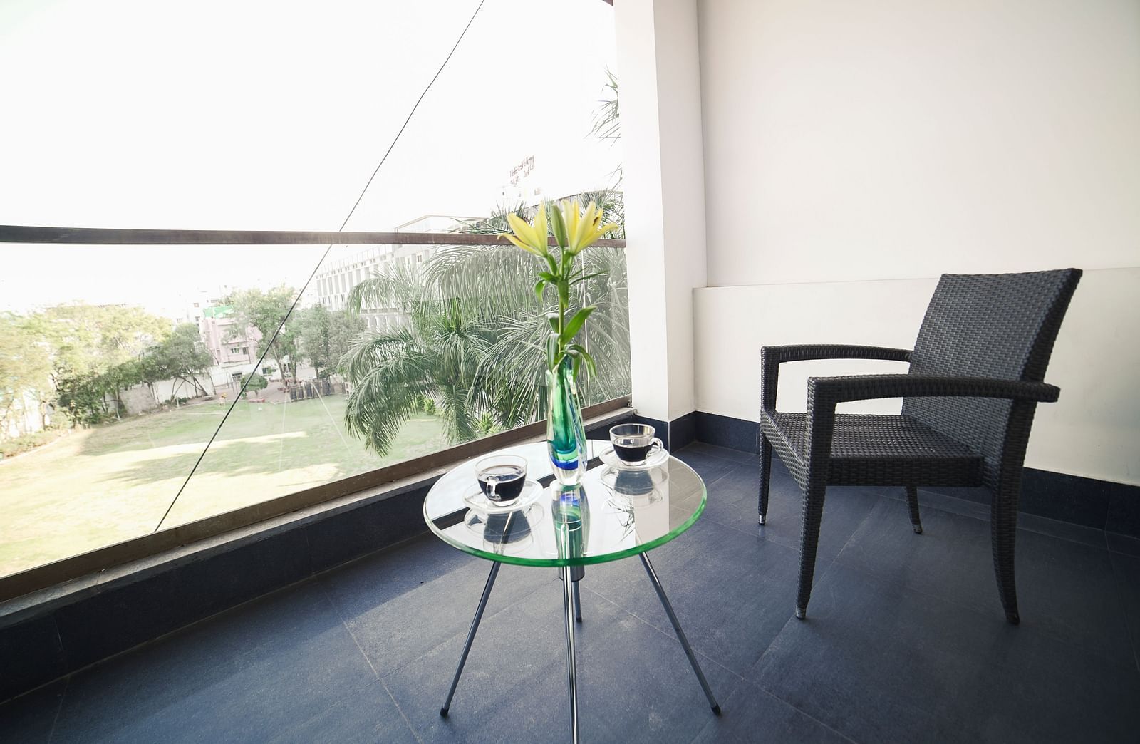 A bright balcony space at BNGV Grandeur Hotel and Banquets with an outdoor chair and a small glass table, offering a view of the surrounding greenery.