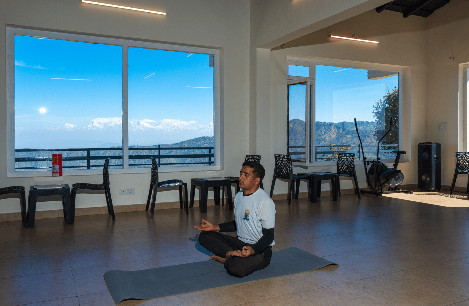 A person sitting in a meditative yoga pose on a mat in a bright studio with large windows at Himalayas Resort By The Lake Hill, Mukteshwar.