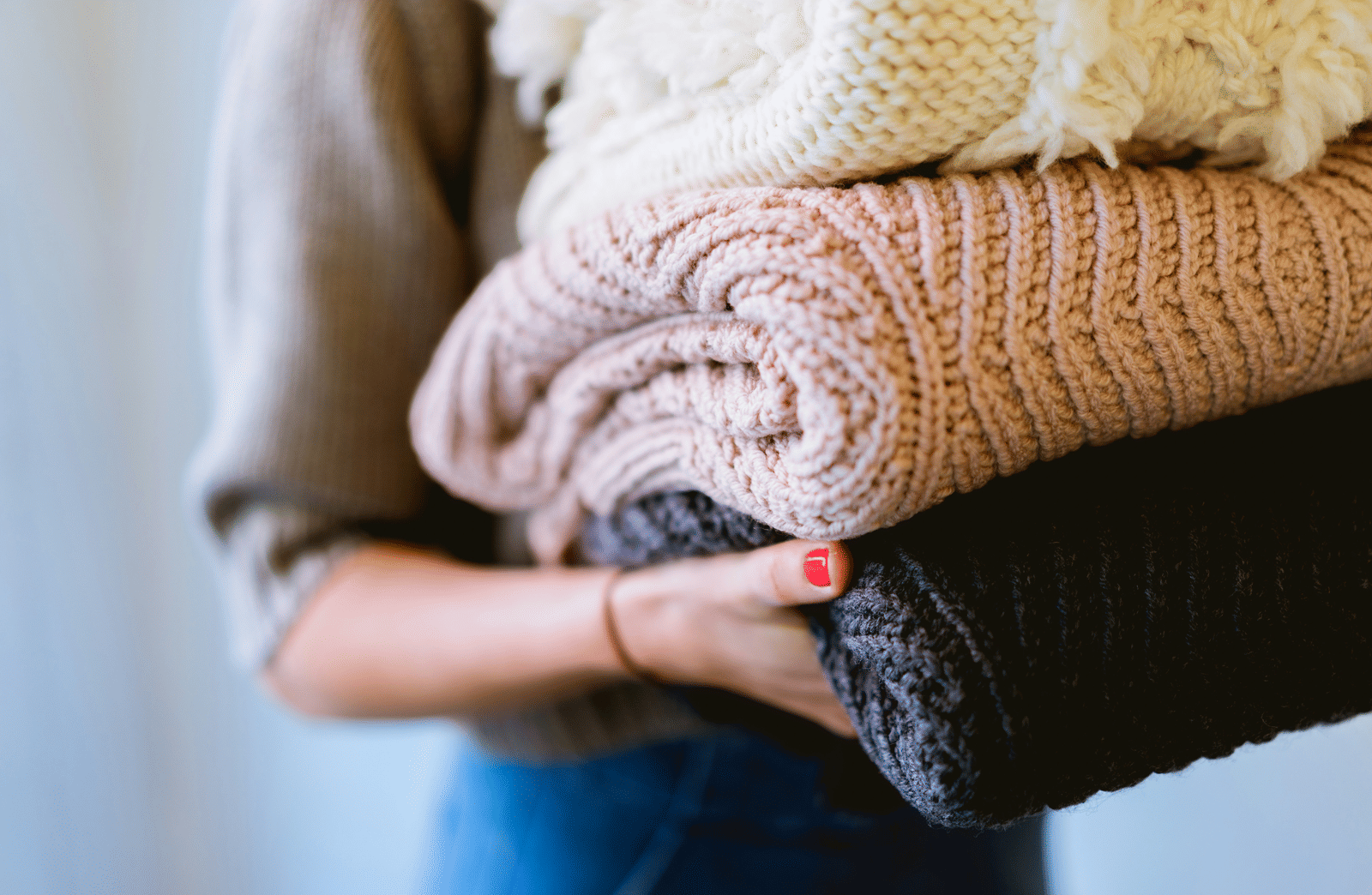 A close-up of a person's hands holding a thick stack of soft, knitted blankets in various neutral shades.