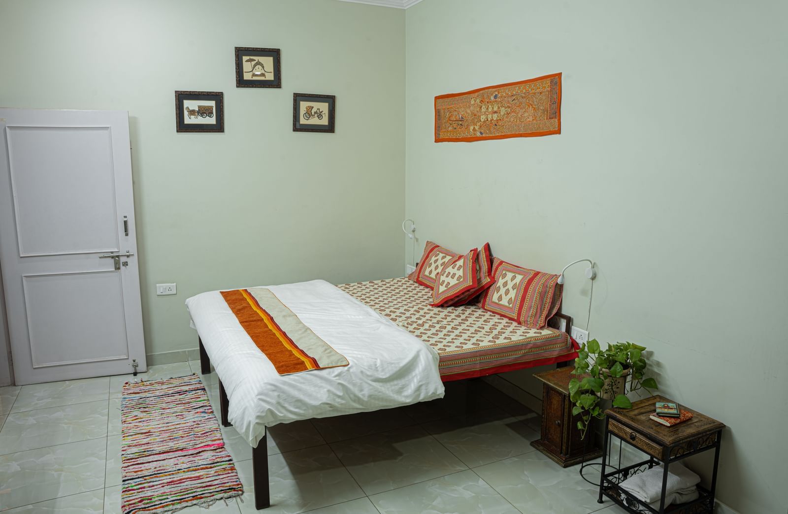 A simple bedroom with a double bed, white bedding, orange throw, wall frames and rug at Jaypore360 Homestay in Jaipur.