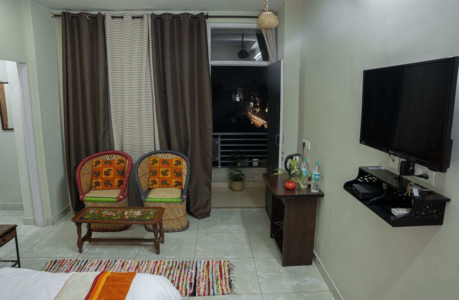 A bedroom with two cushioned chairs, a small table, bed with white bedding and window curtains at Jaypore360 Homestay - Best Rooms in Jaipur.