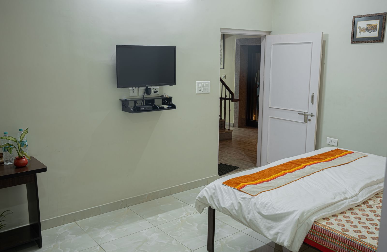 A neatly kept bedroom with a wall-mounted TV, bed, table and doorway visible in the corner at Jaypore360 Homestay - Best Rooms in Jaipur.
