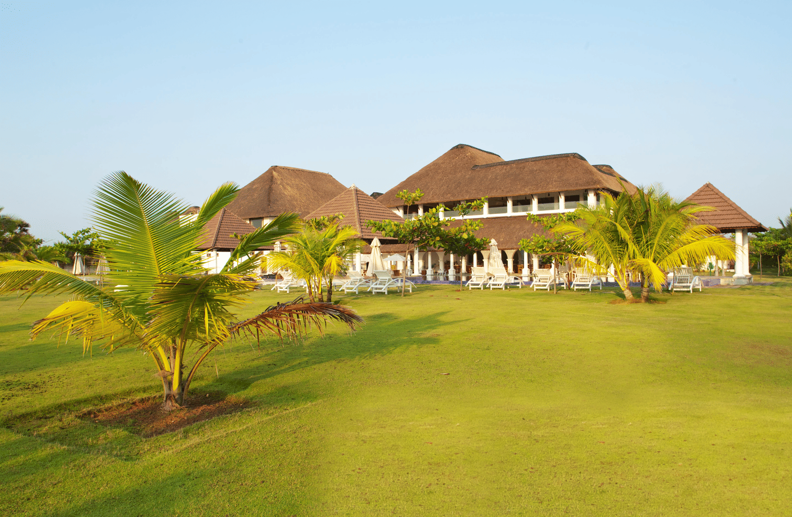 A wide green lawn with small palm trees situated in front of thatched-roof resort buildings | Le Pondy