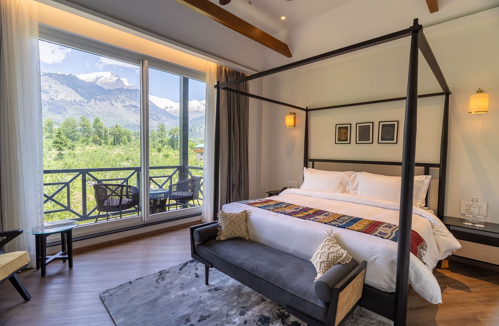 Spacious room with a large wooden bed, cushioned seating at the foot, and a glass door opening to a balcony with a serene nature view at Nivasana Manali.