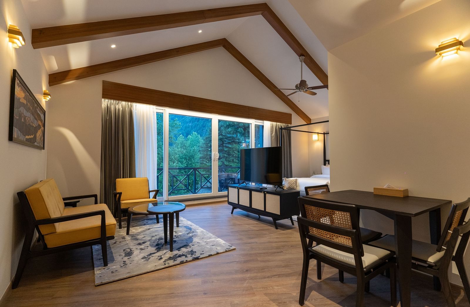 Spacious room with cushioned sofa, wooden table with a TV, and large windows offering scenic nature views at Nivasana Manali.
