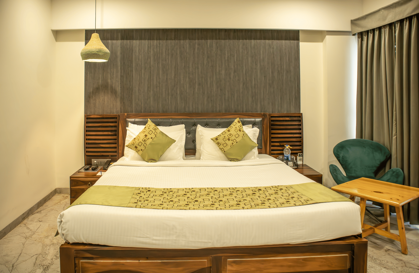 An extended view of a well-lit Premium Deluxe Room at Papaya Tree Hotels featuring a double bed, a small wooden side table with a chair, an overhead lamp, and long curtains.