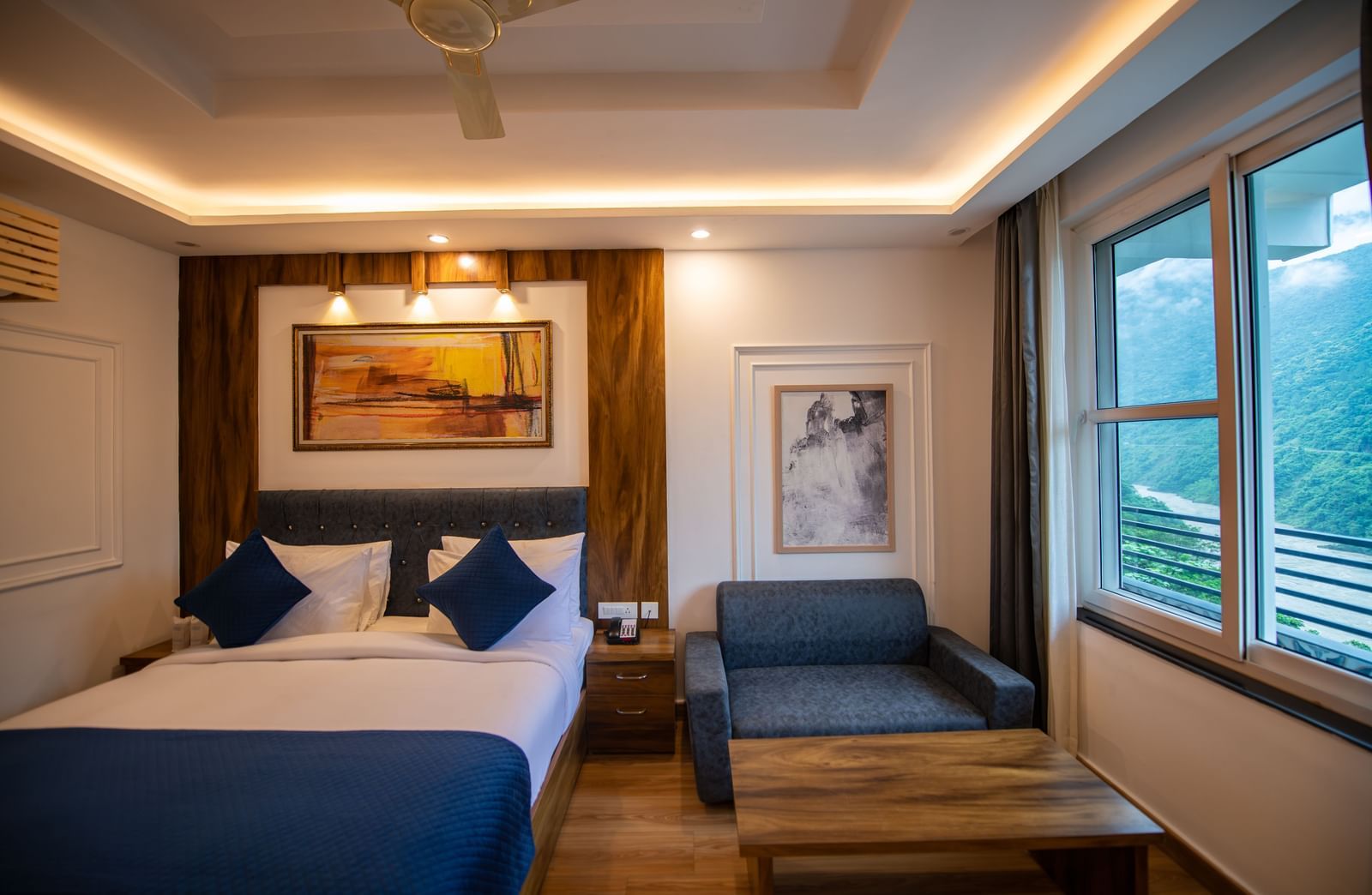 A Deluxe Room at The Tattva Devaprayag with a bed featuring blue blankets and two pillows, two framed paintings on the wall and a sofa.