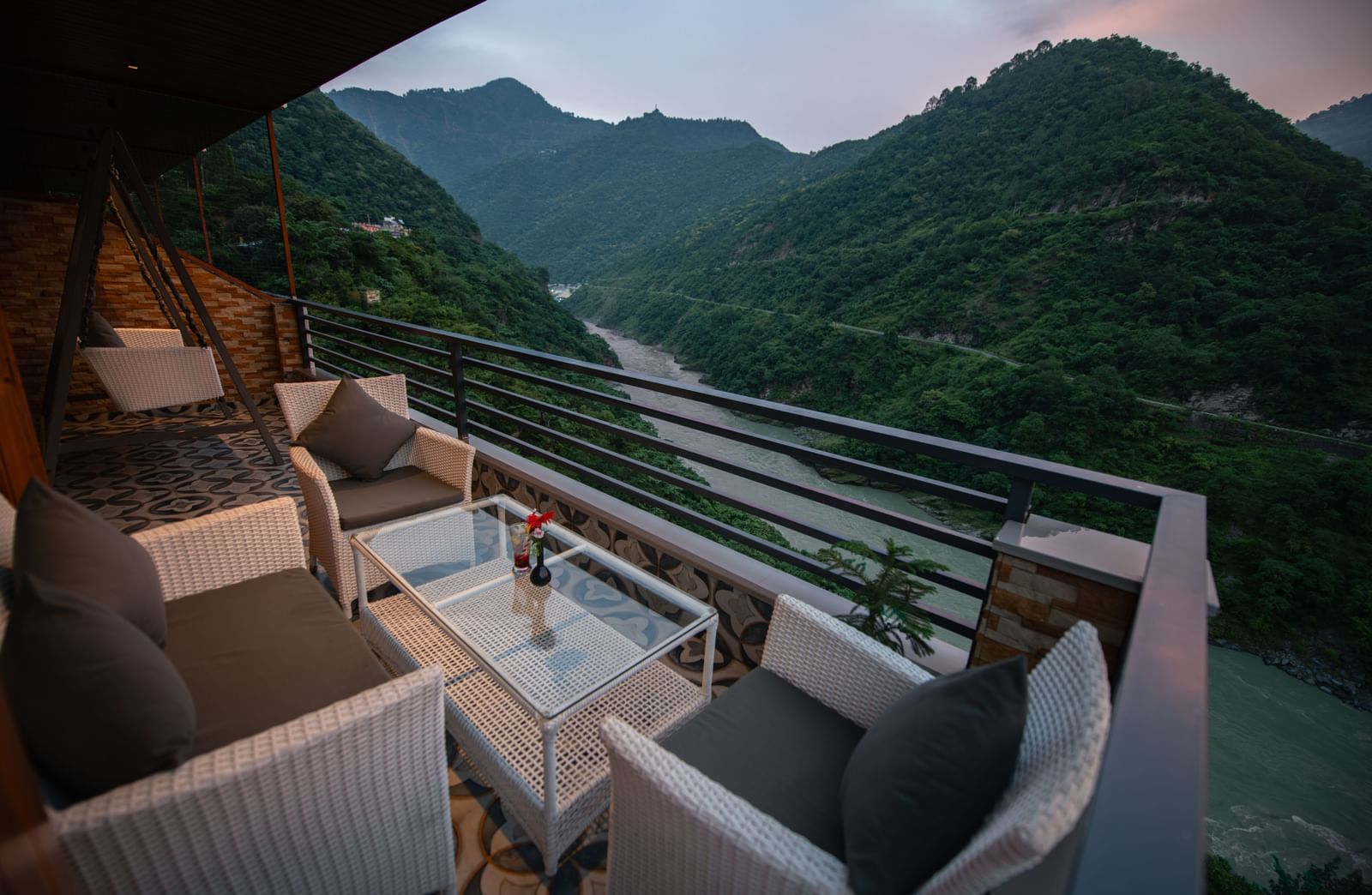 A shot of the lush green mountains and a river flowing by it, as seen from the balcony of the Executive Room at The Tattva Devaprayag featuring a couch and a table.