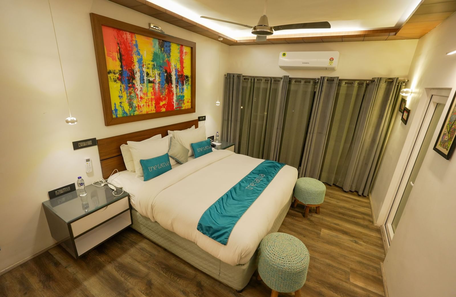 Bedroom of Pent House at The Tattva, Joshimath with a large bed, teal accents, abstract wall art, wooden flooring, grey curtains, and ambient ceiling lighting.