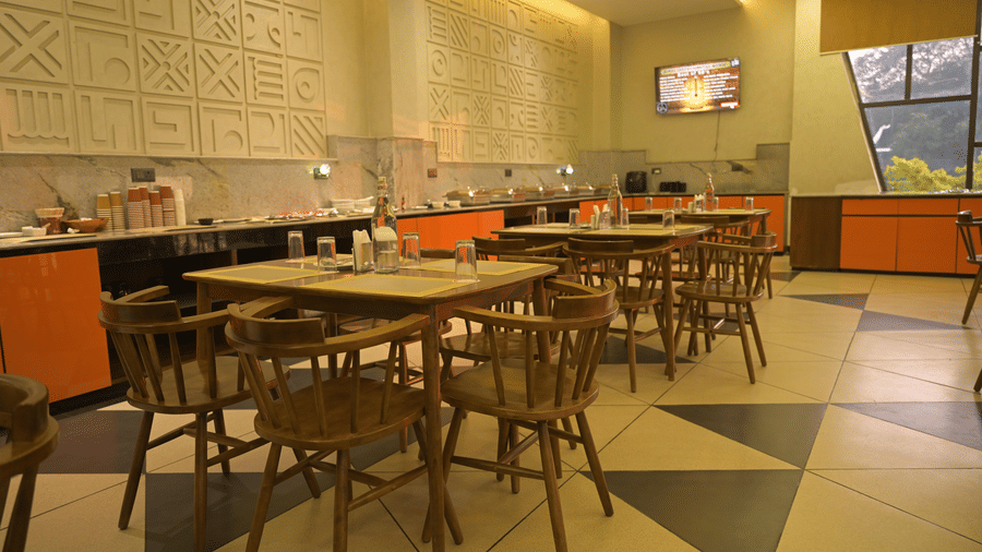 A dining room featuring a wooden table in the foreground and a buffet counter with orange panels in the background at Hotel Sonar Bangla, Mayapur.
