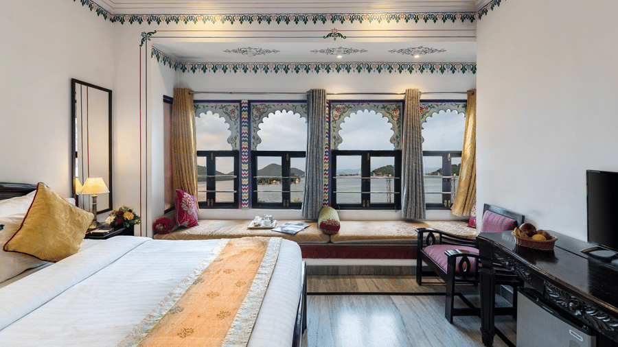 A side view of the bedroom featuring a spacious bed, a wooden table under the wall-mounted television, and bay window seating with curtains pulled to the side at Panna Vilas Palace