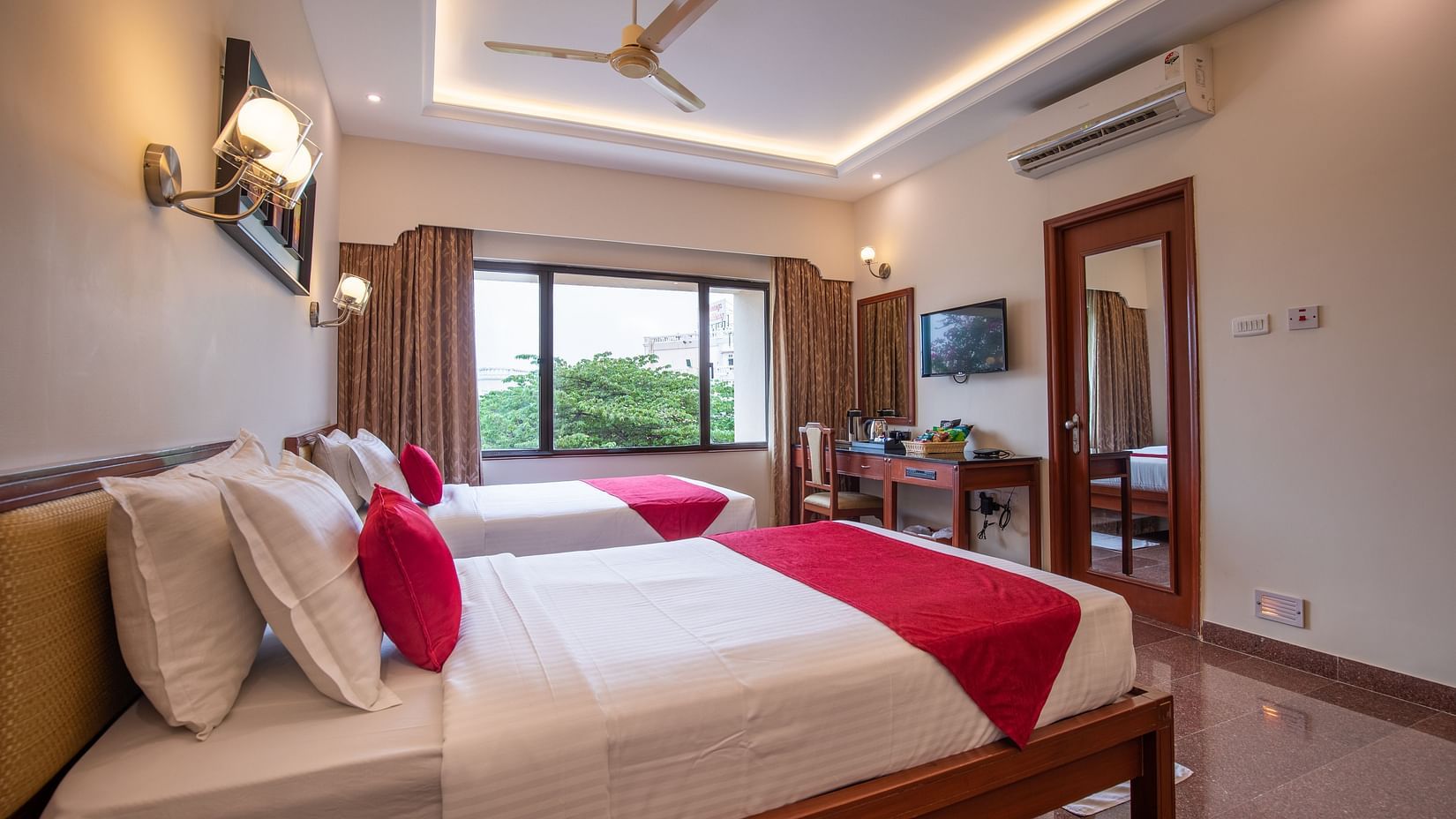 A modern Deluxe Room with twin beds, a large window with curtains, a ceiling fan, TV, desk, and wall-mounted lights