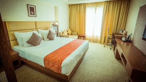 Deluxe room at Pride Hotel and Convention Centre, Indore featuring a plush bed with wooden headboard place in front of a TV mounted on the wall as the light is making through the semi-opaque curtains covering the large window