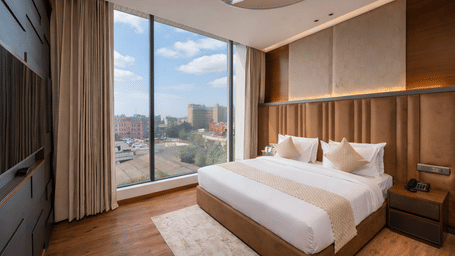   A bedroom with a king size bed, side tables, lamps, curtains, and a window showing buildings outside at DLS Hotels Amritsar.