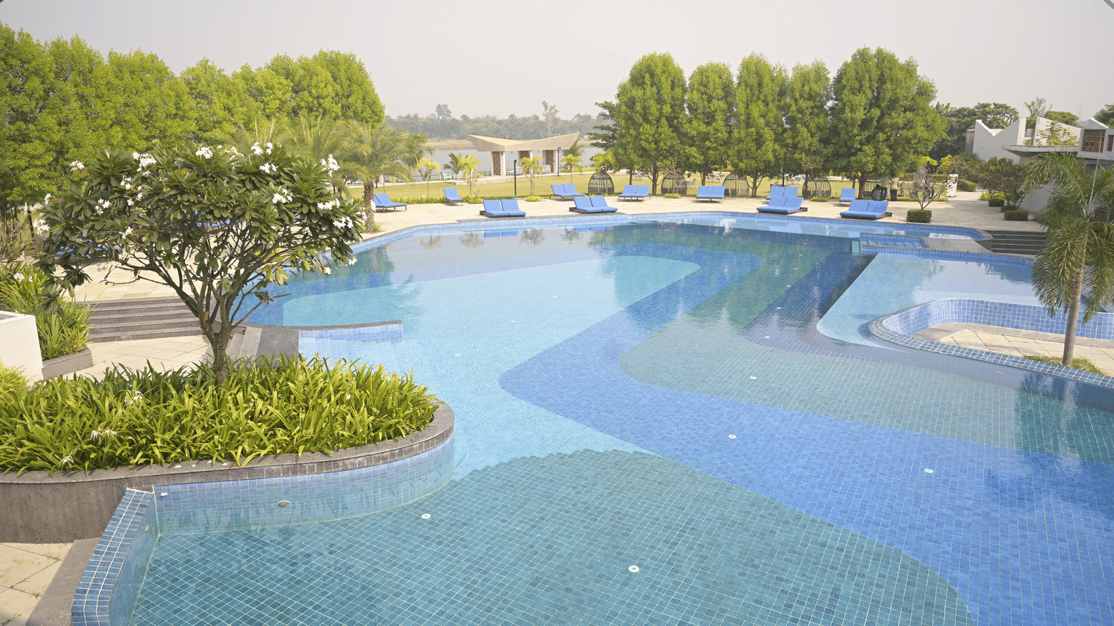 A curved swimming pool surrounded by manicured gardens and open spaces at Hotel Sonar Bangla Mayapur.