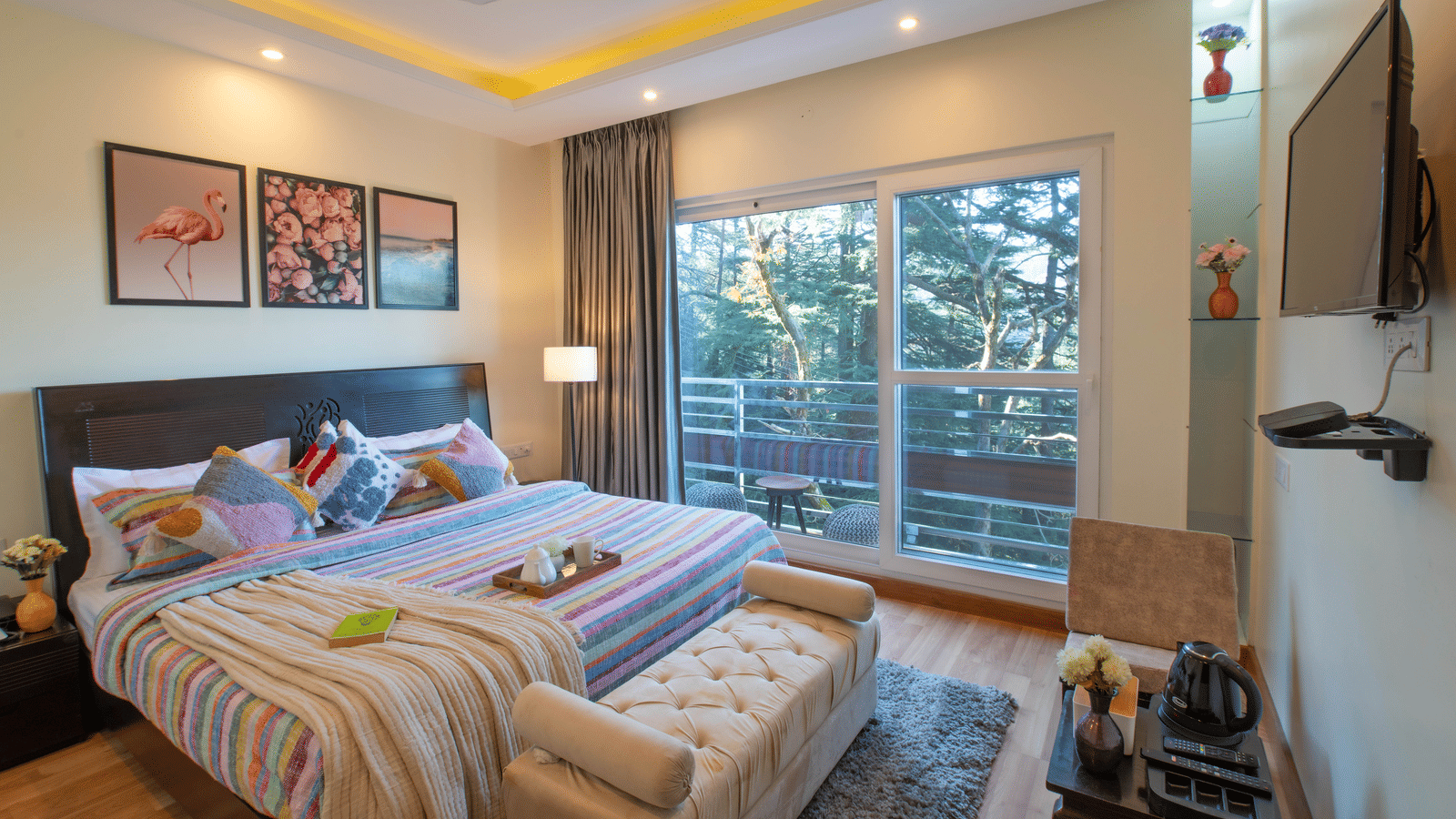 A bedroom at Perfectstayz Value Shimla (Namah Retreat) with a king size bed, wall art, a television, a bench, and glass sliding doors leading to a balcony.