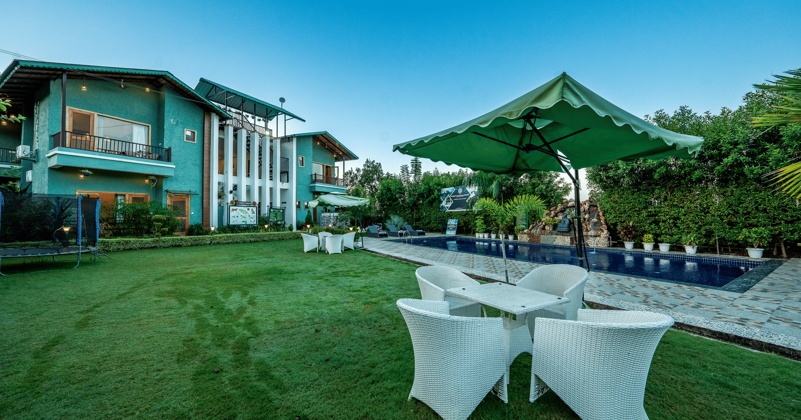 Wide-angle day-time view of the pool area and lawn at Corbett Nirvana Resort, showing the teal-coloured building and white outdoor seating.