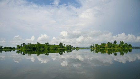 Wide calm river with green tree-lined shoreline reflecting clouds and blue sky, scenic landscape from Tripura, India.