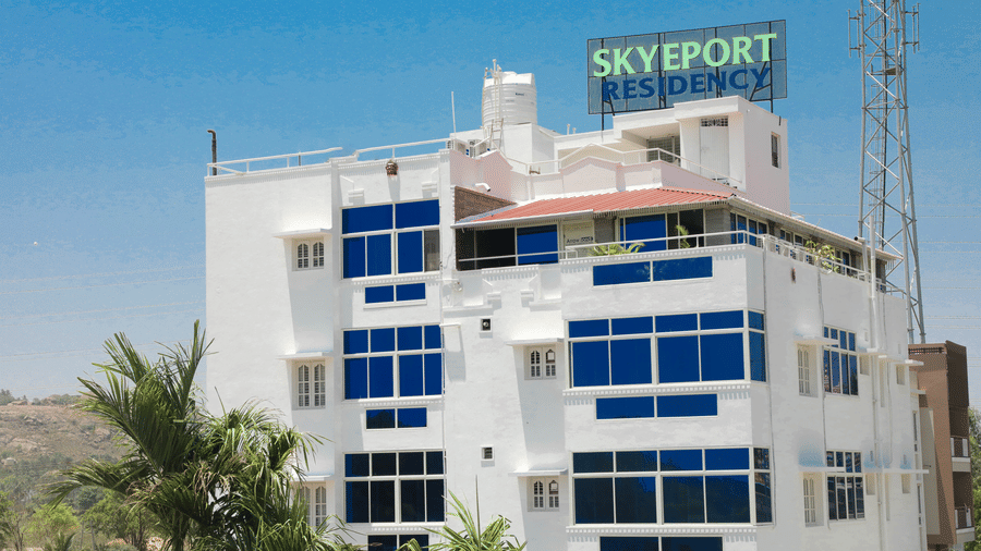 A daytime exterior view of a white multi-storey building with blue glass windows and a sign on top reading Skyeport Residency | Skyeport Residency