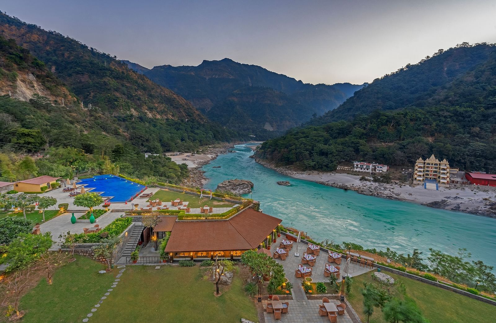 A panoramic view of Aloha on the Ganges, Rishikesh. The image shows the resort nestled amidst hills, with the Ganges River flowing peacefully in the foreground. There is a swimming pool, restaurant, and other facilities visible.
